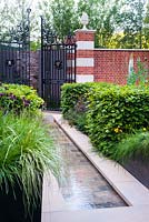 Red brick wall and Bath stone with open ornamental iron gate and a rill paved with Belgian setts slices,  surrounded by Cirsium rivulare 'Trevor's Blue Wonder', Fagus sylvatica hedge, Astrantia 'Hadspen Blood' and grasses - The Living Legacy Garden. RHS Chelsea Flower Show 2015