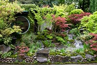 Large round window of house with pinus and acers. Moss on house wall. steps, hard landscaping. Green roof. Waterfall. Edo no Niwa Garden  - RHS Chelsea Flower Show, 2015