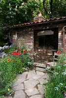 The Old Forge Artisan Garden for Motor Neurone Disease Association - RHS Chelsea Flower Show 2015. Natural rock pathway and vintage brick shed surrounded by Aesculus hippocastanum 'Baumannii', Digitalis purpurea, Papaver rhoeas, Anthriscus sylvestris, Ranunculus acris