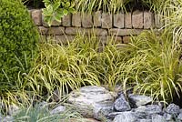 Pavestone Garden - A Garden of Tranquillity. Variegated Grasses with water feature between boulders. Box - Buxus sempervirens to left.