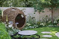 Spherical woven willow bird hide with bench, white flower borders with fencing, circular paving, chamomile lawn and pond with Nymphaea - Living Landscapes: City Twitchers Garden, RHS Hampton Court Palace Flower Show 2015