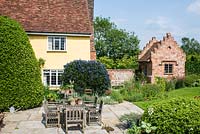 Terrace with wooden table and chairs, terracotta pots.  Laurus nobilis - bay tree, Ceanothus and perennials by a newly built brick garden house with crow stepped gables. Heveningham, June