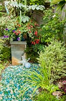 Silver container in border with mixed planting including, Melianthus major, Knautia 'Thunder and Lightening', Petunia, Plectranthus forsteri 'Marginatus', Begonia boliviensis, Buxus, Heuchera and decorative colourful stones and sea shells