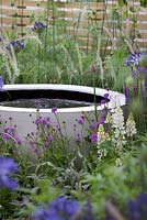 Water feature surrounded by agapanthus, verbena rigida, lupins, salvia and sage. The Wellbeing of Women Garden. RHS Hampton Court Flower Show 2015. Sponsors: Tattersall Landscapes, London Stone, Jacksons Fencing, Hedgeworx, Tactile Studios.  