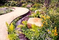Meandering path through garden planted with trees, shrubs, perennials, ferns and large sandstone boulders with adjacent rill in Vestra Wealth: Encore - A Music Lover's Garden at RHS Hampton Court Palace Flower Show 2015 