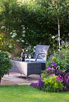 A garden by association, view of seating area with synthetic wicker armchairs and a table surrounded by Betula Jacquemontii, Anthriscus sylvestris, flowerbed with Cosmos,  Veronica longifolia 'Charlotte', Nepeta x faassenii 'Blue Wonder', Sambucus nigra porphyrophylla 'Eva' and pot with Pittosporum Tobira. Designer: Tina Vallis, MSGD