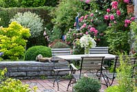 Wood and iron garden furniture on a brick stone terrace in a pottery maker's garden with ceramic objects and Rosa 'Leonardo da Vinci', 'New Dawn', 'Rosarium Uetersen', Acer japonicum 'Aconitifolium', Buxus and Sarracenia