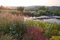 Sunloungers on deck next to pond with Penstemon, Silene, Eryngium - Sea Holly, Euphorbia, grasses Butomus umbellatus -flowering rush at dawn. Follers Manor, Sussex. Designed by: Ian Kitson