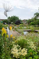 View across pond with stainless steel Seedhead by Ian Marlow ARBS, surrounded by Anthemis tinctoria 'E.C Buxton', Kniphofia 'Green Jade' and Knautia macedonica - The Walled Garden at Mells, Somerset