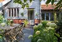 View of shady terraced area with wooden table and chairs. Hydrangea arborescens 'Annabelle' in border 