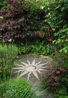 Sun mosaic in secluded corner of garden. Cercis canadensis 'Forest Pansy', roses, rosemary, Berberis thunbergii f. atropurpurea 'Rose Glow'