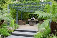 Steps leading up to pergola with Vinca minor 'Alba' and Polystichum polyblepharum, outdoor wood fired oven on table made from wire cage filled with pebbles, borders include Dicksonia antarctica, Cenolophium denudatum and Tiarella  - Live Outdoors, RHS Hampton Court Palace Flower Show 2012