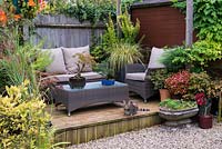 Tucked away in quiet corner at bottom of garden, between shed and greenhouse, a raised wooden deck with outdoor rattan furniture and tabby cat Trixie. Pots with bonsai trees, coleus and ornamental grasses.