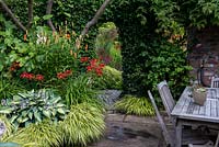 An ivy clad brick wall with door leadind to an herbaceous area. In the foreground the border is planted with Hostas, Hakonechloa grasses, red daylilies and orange Lilium martagon.