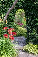 An arch in an ivy clad brick wall leading to an herbaceous area. In the foreground the border is planted with Hakonechloa grasses, red daylilies and orange Lilium martagon.