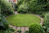 A town garden with circular lawn surrounded by a stone path and foliage plants including Buxus sempervirens balls and Cornus alba 'Elegantissima' shrubs.