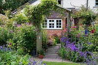 A cottage garden with wooden arch surrounded by Geranium x magnificum, G. psilostemon and G. 'Rozanne' with roses and clematis.