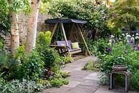 In left bed, silver birch with face sculpture by Pauline Lee amidst euphorbia, black elder, perennial wallflower, grasses and aquilegias. A swing seat sits below pink camellias and Rosa 'Paul's Himalayan Musk'. On right, sunny bed planted with purple and white alliums, scabious, peonies, Mathiasella burpleuroides 'Green Dream' and Iris 'Jane Phillips'.