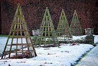 Herbaceous borders covered in snow with wooden pyramid obelisks at Levens Hall and Garden, Cumbria, UK. 