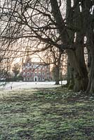 Dawn sunlight rises above the early C18th Queen Anne facade of Welford House illuminating the frosty ground dotted with snowdrops and aconites. Welford Park, Newbury, Berks, UK