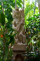 A Balinese carved stone statue of Saraswati a Hindu deity, playing a stringed instrument on a carved plinth in a shady garden surrounded by lush plants.