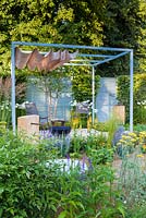 Retreat Garden, Steel pergola with canvas awning stands over metal table and chairs on white paved raised terrace area with gravel surrounding. A circular water feature sits in front. Planting is direct into the gravel, with drought tolerant plants including verbascum, achillea, echinacea, artemisia, geum, perovskia and Nandina domestica. RHS Hampton Court Flower Show in 2016