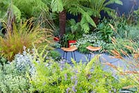 The Bowel Disease UK Garden for Crohn's Disease, view of two padded seats on grey curved paving. Plants in the foreground: Duranta 'Geisha Girl', Artemisia ludoviciana 'Silver Queen', Halimocistus wintonensis and Baloskion tetraphyllum. Pseudopanax crassifolium and tree ferns - Dicksonia antarctica and D. squarrosa behind, with Hosta 'Patriot', Canna indica, Pinus strobus nana radiata and Westringia fruticosa. RHS Hampton Court Flower Show in 2016