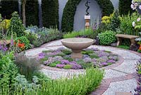 The St John's Hospice Garden, The Modern Apothecary. Extensive planting of herbs around water basin in central circular bed with cobble stone surround. oak seat in border and sculpture of Aesclepius on back wall.  RHS chelsea Flower show 2016