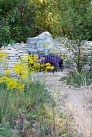 L'Occitan Garden, view of earth path leading to a patio with black metal chairs, table with purple table cloth surrounded by Galium verum, Juglans regia. The RHS Chelsea Flower Show 2016, Designer: James Basson MSGD, Sponsor: L'Occitane en Provence