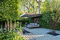 The Husqvarna Garden, view of modern patio seating area with furniture, bronze sculpture, bluestone cobbled paths, surrounded by  Digitalis purpurea, Prunus maackii 'Amber Beauty', Carpinus pleached floating cubes, Taxus hedging. RHS Chelsea Flower Show, 2016.