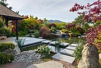 A garden building and stepping stones over a pond with Pinus sylvestris and Acer palmatum  trees, A Japanese Reflection, RHS Malvern Spring Festival 2016. Design: Peter Dowle and Richard Jasper, Howle Hill Nursery