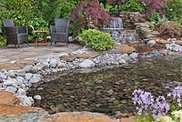 A patio with natural stone slabs and chairs by a naturalistic pool fed by a waterfall. The Water Garden, RHS Tatton Flower Show 2011, Cheshire