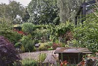 A view across a patio seating area and gravel pathway to a bridge over a small pond surrounded by planting of mixed perennials, shrubs and trees, Cheshire