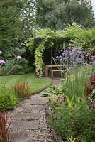 A gravel and stone slab path between flower borders and lawn leading to a shady arbour seating area with climbing plants, Cheshire