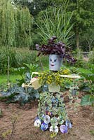 A scarecrow made from galvanised buckets, compact discs, wood and string. Plants included are Variegated Helichrysum, Iresine, Sanvitalia procumbens 'Sunvy Trailing' and Panicum virgatum 'Warrior'