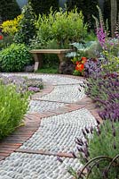 A Modern Apothecary. A brick and cobble path leading to an oak bench is edged in French lavender, Lavandula stoechas 'Helmsdale', and Lavandula viridis 'Jekka's Blue'. Designer: Jekka McVicar. RHS Chelsea Flower Show 2016