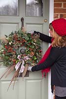 Sheree King hanging a traditional christmas wreath on a front door. Featuring Eucalyptus gunnii, Rosa 'Bonica' rose hips, Variegated Ivy, Ilex aquifolium, Cotoneaster lacteus, Pinus nobilis, Godetia and Miscanthus sinensis seed heads