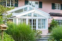A glasshouse porch flanked with ornamental glasses joins house and garden, Miscanthus sinensis 'Gracillimus'