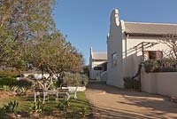 White painted and dark wood chairs and table next to building with sand paths, borders with Agave attenuata and landscape view beyond - August, Naries Namakwa Retreat, Namaqualand, South Africa