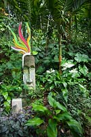 A lush thickly planted garden featuring a white flowering, Eucharis grandiflora, Eucharist lily and a carved stone head decorated with a brightly painted panel of hand painted glass.