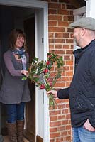 A man handing a rustic Valentine's wreath to his partner. Made with Hedera helix, Willow stems, Birch twigs, Cornus sanguinea 'Midwinter Fire', Tulip 'Rococo', Viburnum tinus and Chamelaucium