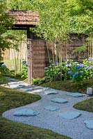 View of the path with slate paving stones and entrance to the garden surrounded by Phyllostachys bissetii, blue flowering Hydrangea and knitted Bamboo fence. Japanese Summer Garden. Designed by Saori Imoto. Sponsored by Unique Japan Tours