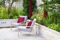 A pair of grey padded stainless steel Barlow Tyrie Equinox' Armchairs with burgundy cushions and pergola on Yorkstone paved terrace, surrounded by raised beds. Plants include Achillea millefolium 'Paprika', Pennisetum orientale, Penstemon 'Port Wine', Sanguisorba 'Chocolate Tip' and Taxus baccata.  Squire's 80th Anniversary Garden, RHS Hampton Court Flower Show 2016. Designer: Catherine MacDonald - Sponsor: Squire's Garden Centres