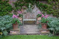 Decorative ironwork seat on stone patio flanked  with terracotta pots filled with Tulipa 'China Pink' underplanted with a dark red Pansy,and  silver foliage of Artichokes  in the potager.Pashley Manor