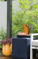 Detail of a white outdoor lounge with a blue cube table with an orange metal pitcher, orange patterned drinking glasses an yellow plastic pot planted with an Alternanthera ficoidea with burgundy red foliage in front of a courtyard wall with a Bambusa textilis 'Gracilis'.