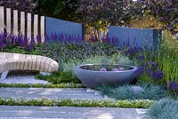 Gravel and stone patio with curved bleached oak wooden bench and fence, grey rendered wall, concrete containers and small pond. Planting includes Thyme and Rosemary, Salvia officinalis 'Purpurascens', Salvia tanzerin, Perovskia atriplicifolia 'Blue Spire'.  Living Landscapes: Healing Urban Garden, RHS Hampton Court Palace Flower Show 2015. Designer Rae Wilkinson