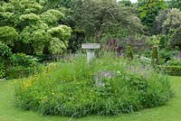 A cottage garden with circular lawn and raised wildflower mound planted with buttercups, cranesbill, cow parsley. At the centre, a raised stone bird bath.