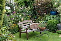 In grassy corner of small suburban garden, bench and table with pot of diascia. Behind, bed of Japanese anemones, cleome, rudbeckia and roses.