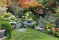In grassy corner of small suburban garden,  seating area edged by bed of hostas, acer, hydrangea and box.