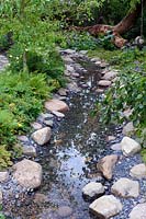 The Zoflora and Caudwell Children's Wild Garden. Shallow stream in woodland garden with edging of rocks and pebbles. Designers: Adam White and Andree Davies. Sponsors: Zoflora. RHS Hampton Court Palace Flower Show 2017